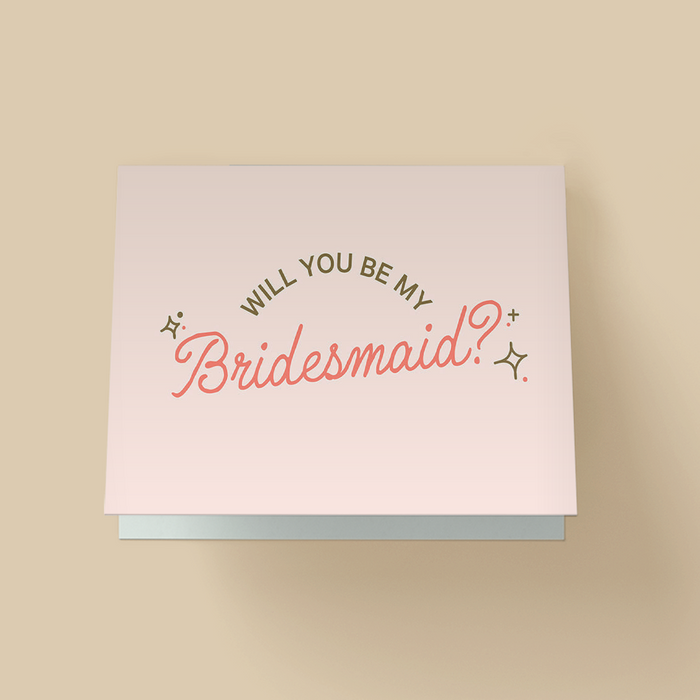 White bridesmaid proposal card. Small uppercase sans-serif text "WILL YOU BE MY" arched on top, and large coral-pink cursive "Bridesmaid?" below, with small star sparkle accents on the sides.