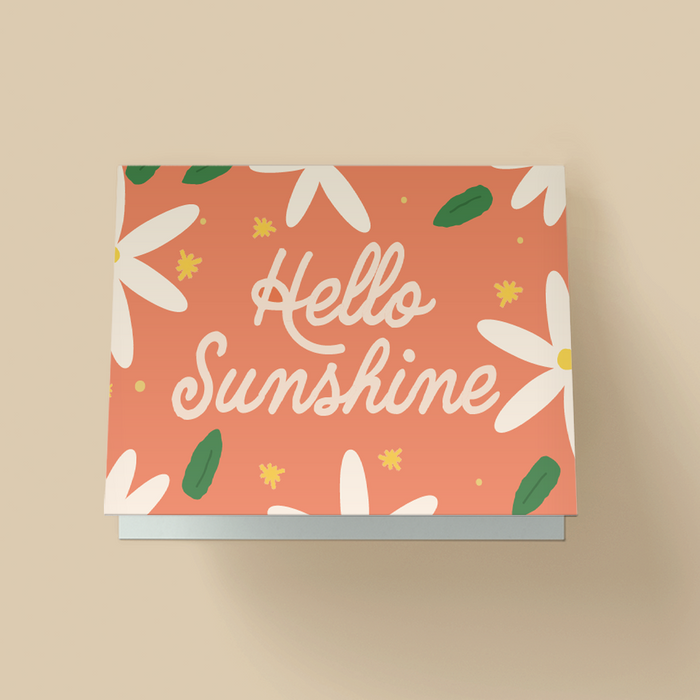 Greeting card with a solid coral-orange background covered in a floral pattern of large white daises, green leaves and small yellow stars. The word "Hello Sunshine" are written in large, white, playful cursive script in the center