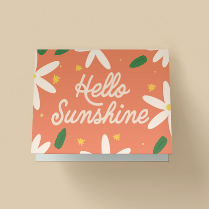Greeting card with a solid coral-orange background covered in a floral pattern of large white daises, green leaves and small yellow stars. The word "Hello Sunshine" are written in large, white, playful cursive script in the center