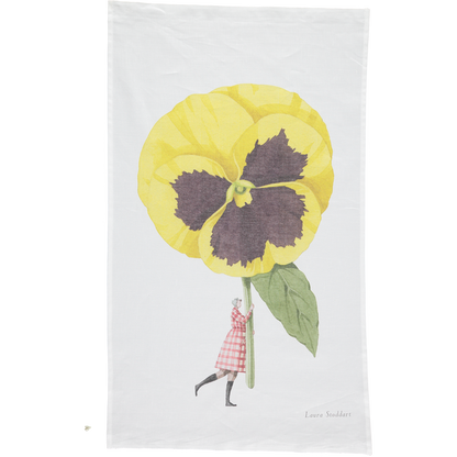 A white tea towel featuring a whimsical illustration of a small woman holding a gigantic yelllow pansy by the stem. Art by Laura Stoddart.