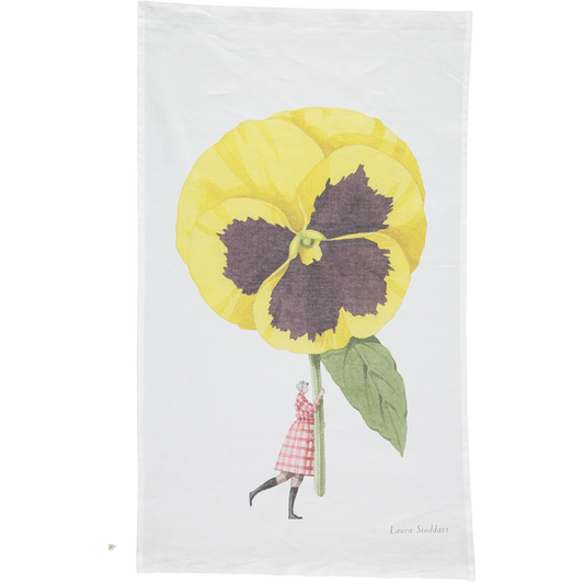 A white tea towel featuring a whimsical illustration of a small woman holding a gigantic yelllow pansy by the stem. Art by Laura Stoddart.
