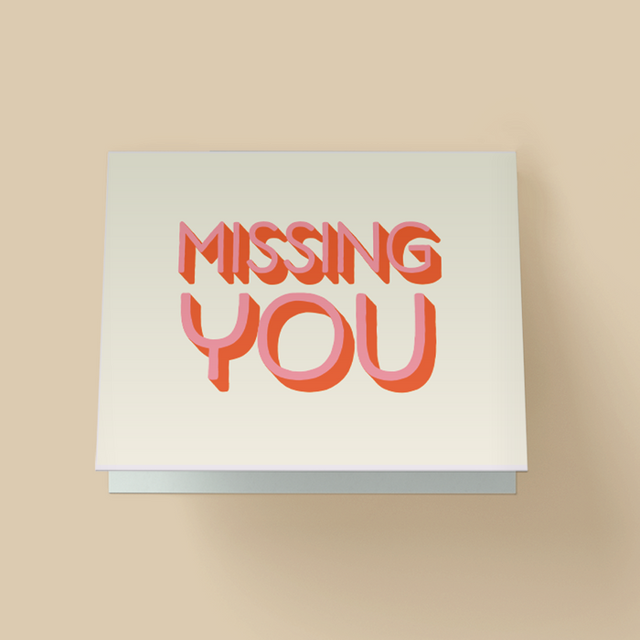 White greeting card. The words "MISSING YOU" are in large, retro-style #D block letters - light pink font face with burnt orange shadow/extrusion - stacked in two lines and centered.