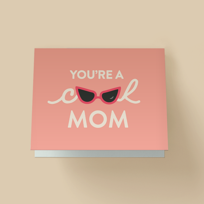 Mother's Day greeting card with a solid peach background. White text reads ' YOU'RE A cool MOM' - 'cool' is in cursive script, with red cat-eye sunglasses replacing the double O. The sunglasses have black lenses
