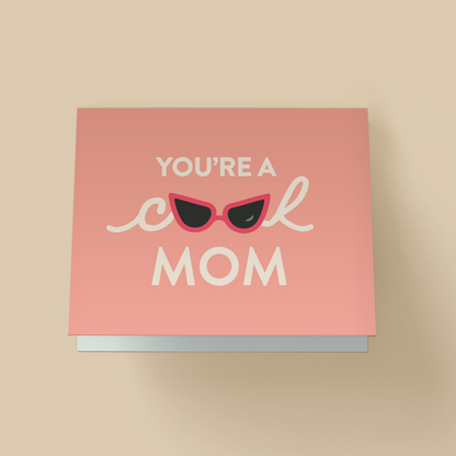 Mother's Day greeting card with a solid peach background. White text reads ' YOU'RE A cool MOM' - 'cool' is in cursive script, with red cat-eye sunglasses replacing the double O. The sunglasses have black lenses